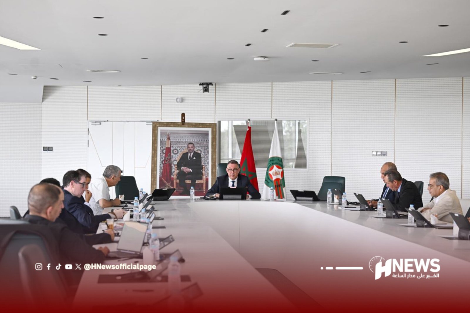 A formal meeting in a bright conference room with many attendees seated around a long table, a man in a suit sits at the center near flags and a portrait backdrop.