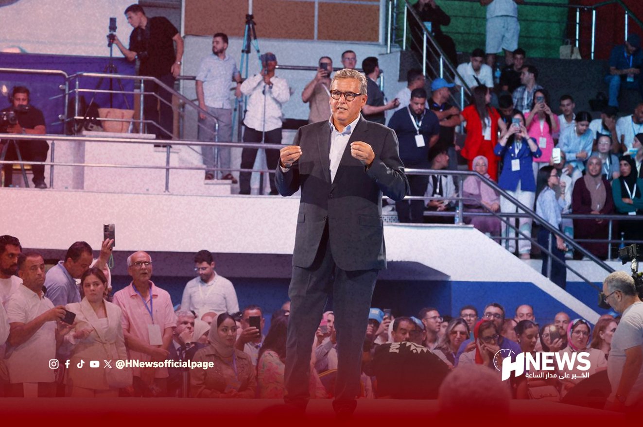 A man in a dark suit speaks on a stage to a large crowd at a news event, with cameras and reporters behind him; HNews logo bottom right.