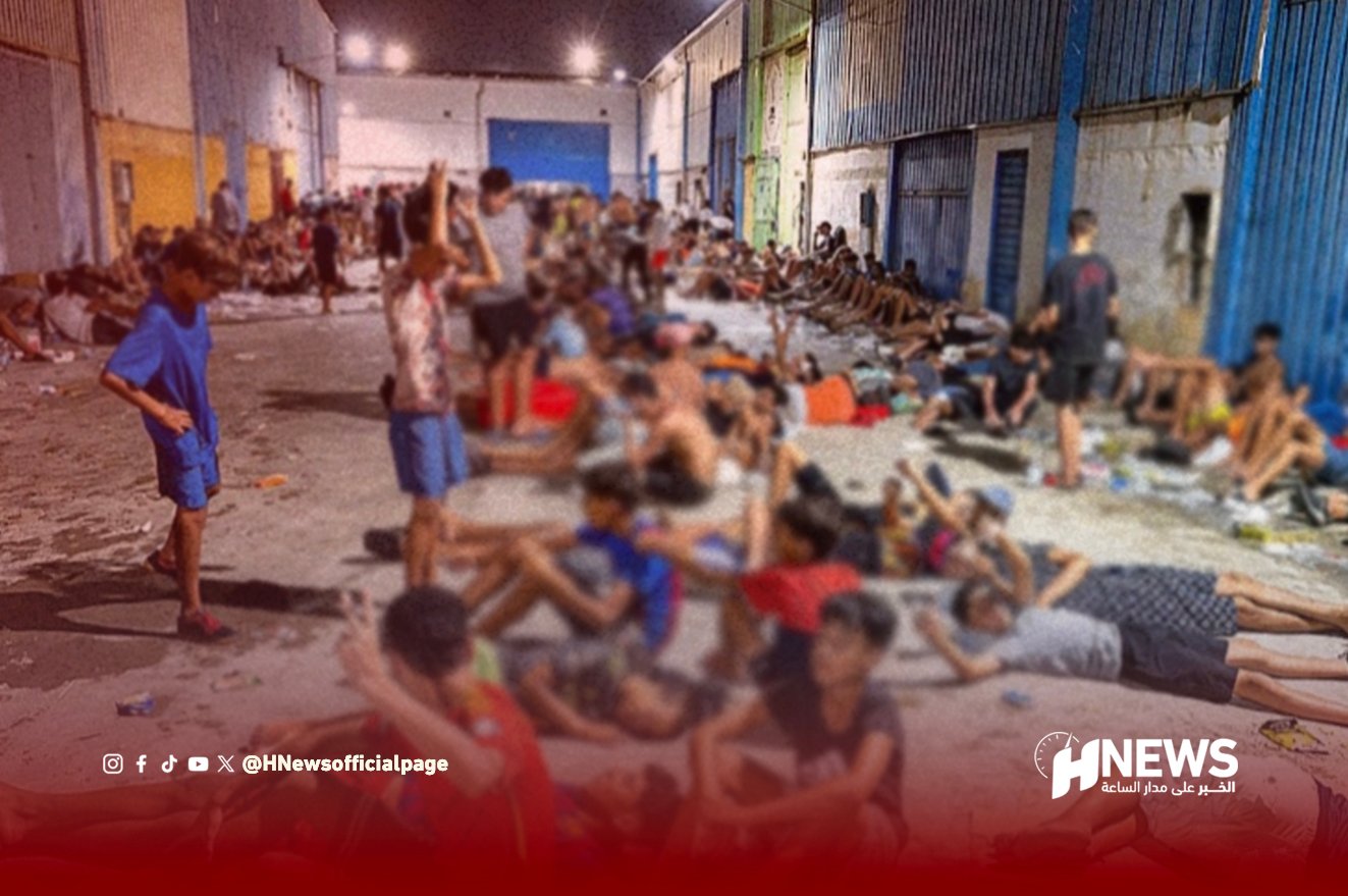 Crowded indoor shelter with people sitting and lying on the floor along a long hallway, appearing tired in a humanitarian setting.