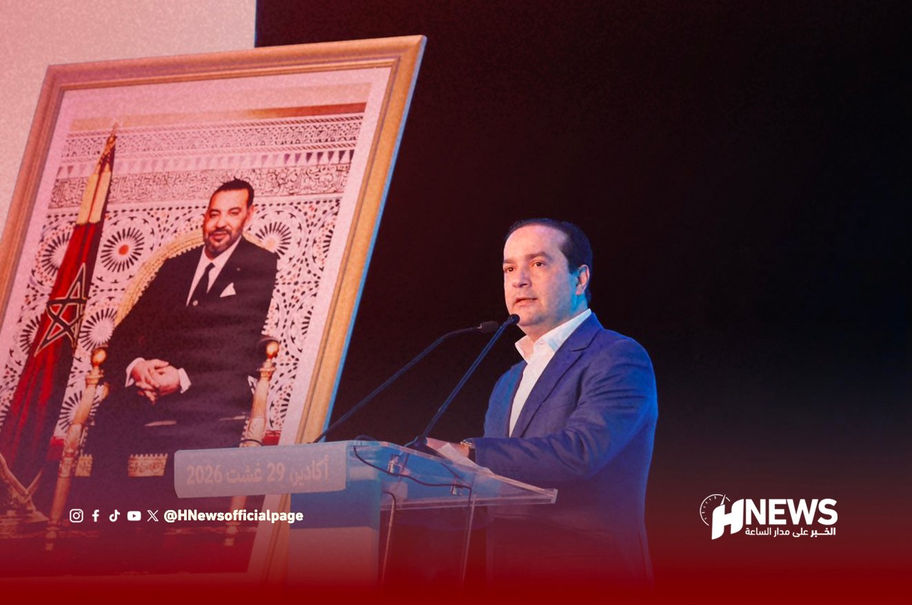 Man in a blue suit speaks at a clear podium on a stage, with a large framed portrait and flag visible to his left.