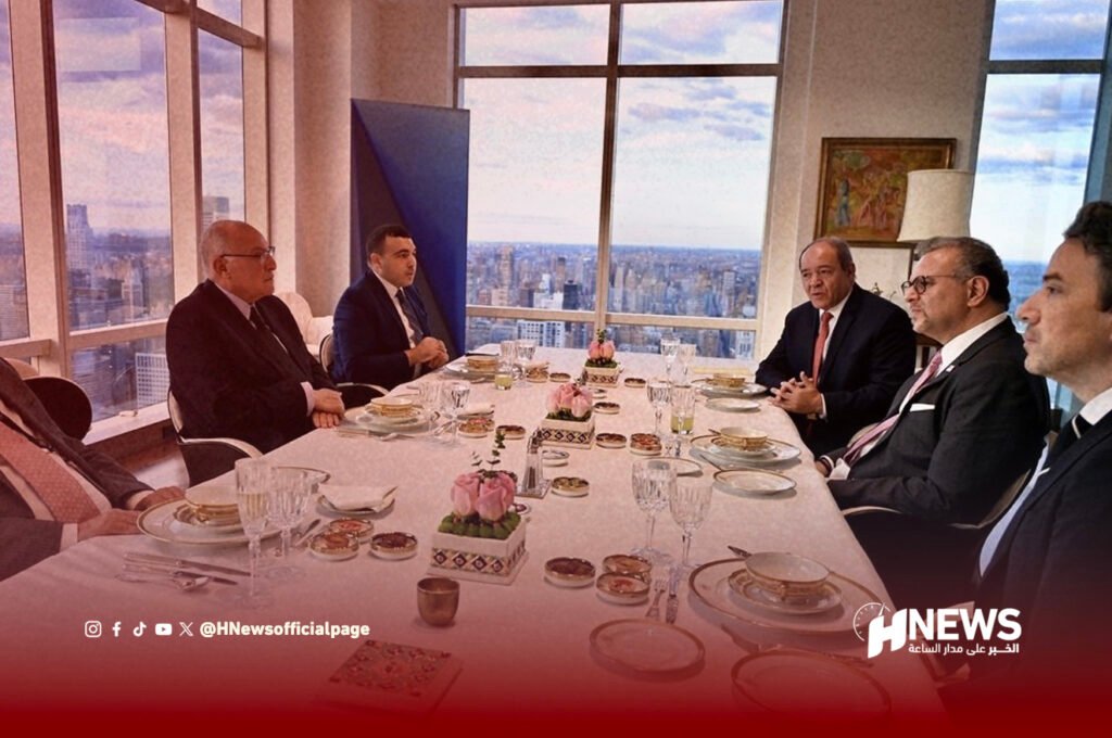 بولس وعطاف | H-NEWS آش نيوز Business professionals in suits sit around a long dining table in a high-rise restaurant with a city skyline in the background, floral centerpieces present.
