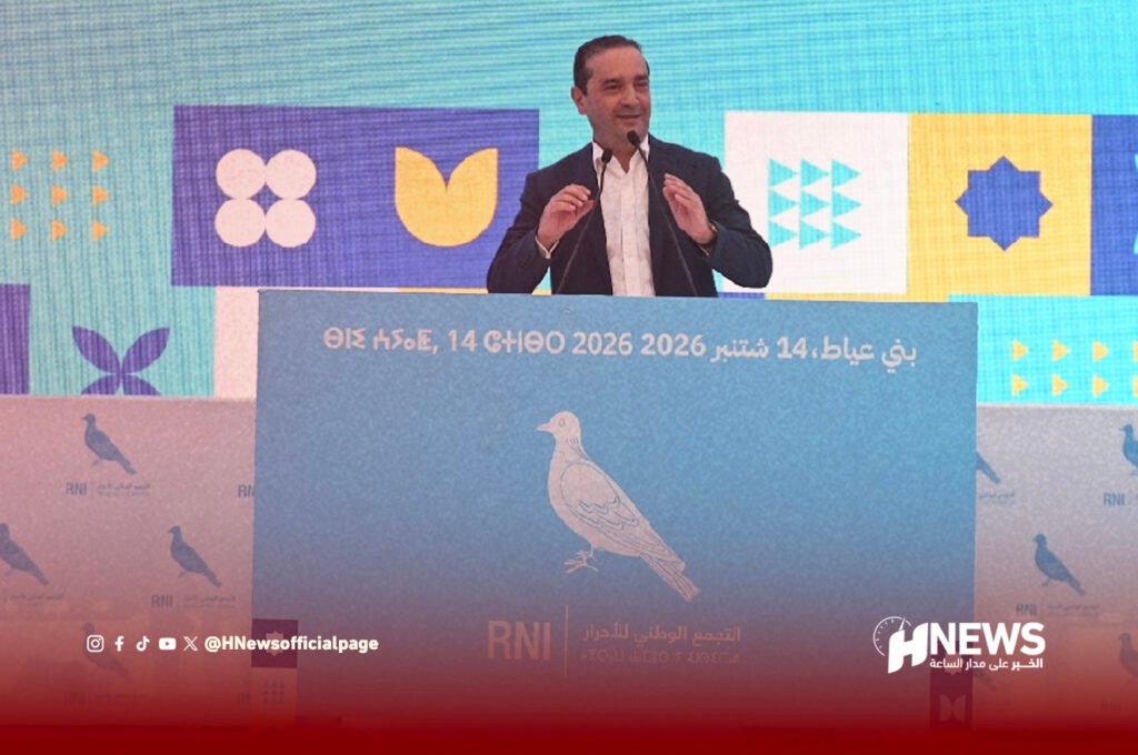 Speaker in a suit delivers a speech at a podium on a blue backdrop with Arabic text and geometric shapes in the background.