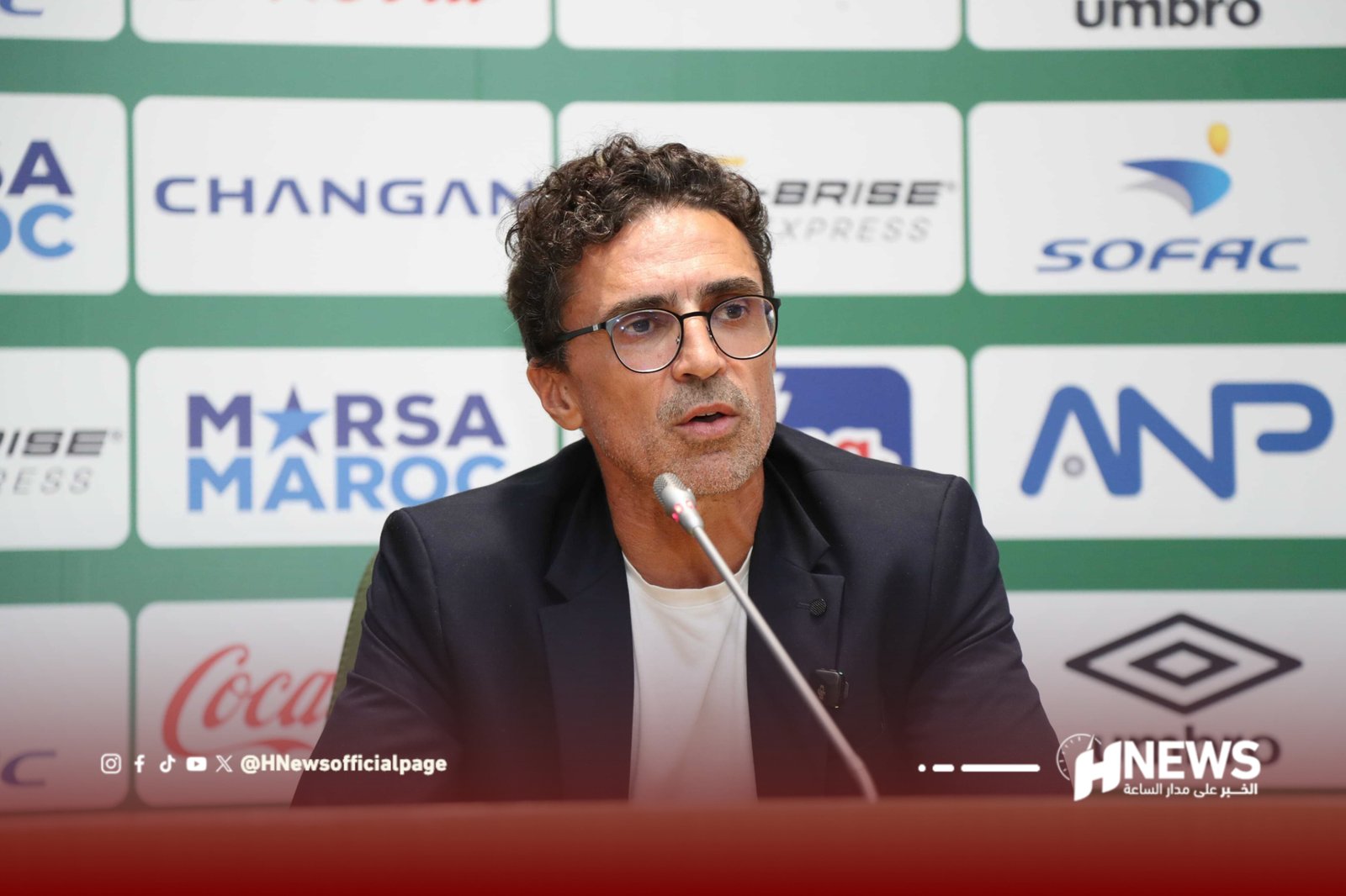 Man with glasses speaking at a press conference, seated in front of a green sponsor backdrop and microphone in frame