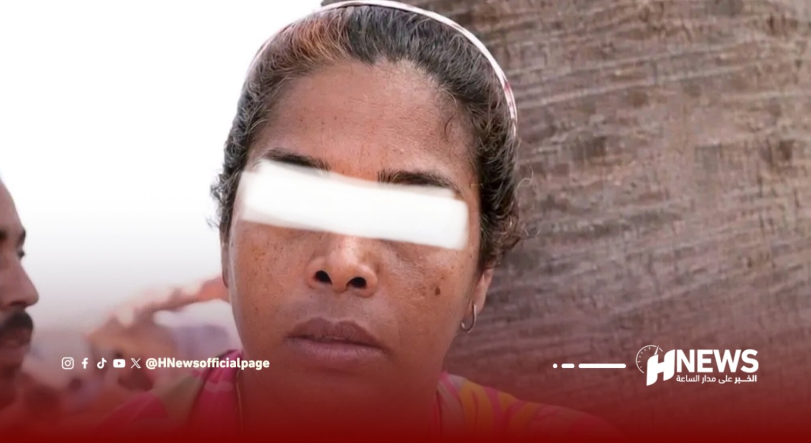 Woman with a white band covering her eyes, facing the camera; brick wall behind and a news watermark in the bottom corner.