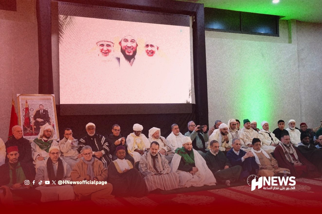 Group of men in traditional clothing seated on a carpeted floor at a ceremony, with a large screen showing three smiling faces behind them and a framed portrait nearby.
