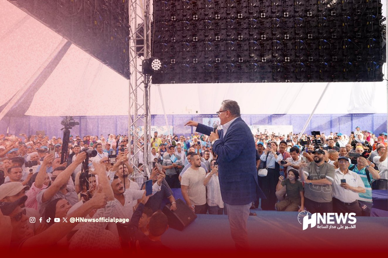 Speaker wearing a blue blazer addresses a large crowd on a stage at an outdoor event, with a news logo in the corner and red wash at bottom.