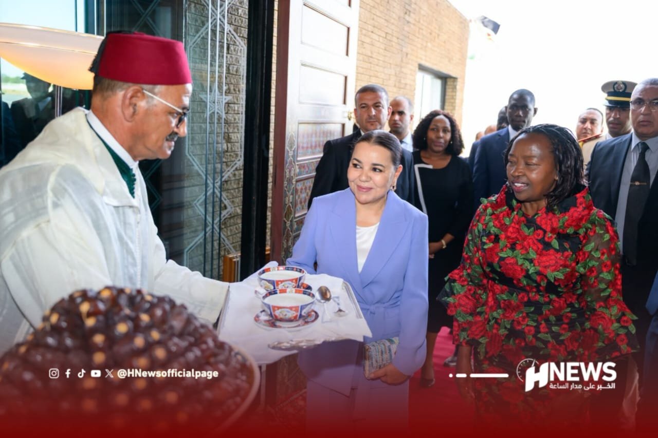 Official-looking man in a red fez serves tea to a smiling woman in a lavender suit at a doorway, with a crowd watching.