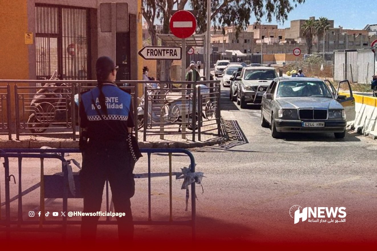 Border crossing scene: security officer in a blue vest stands behind barriers as cars queue at a checkpoint with a 'Frontera' sign visible.