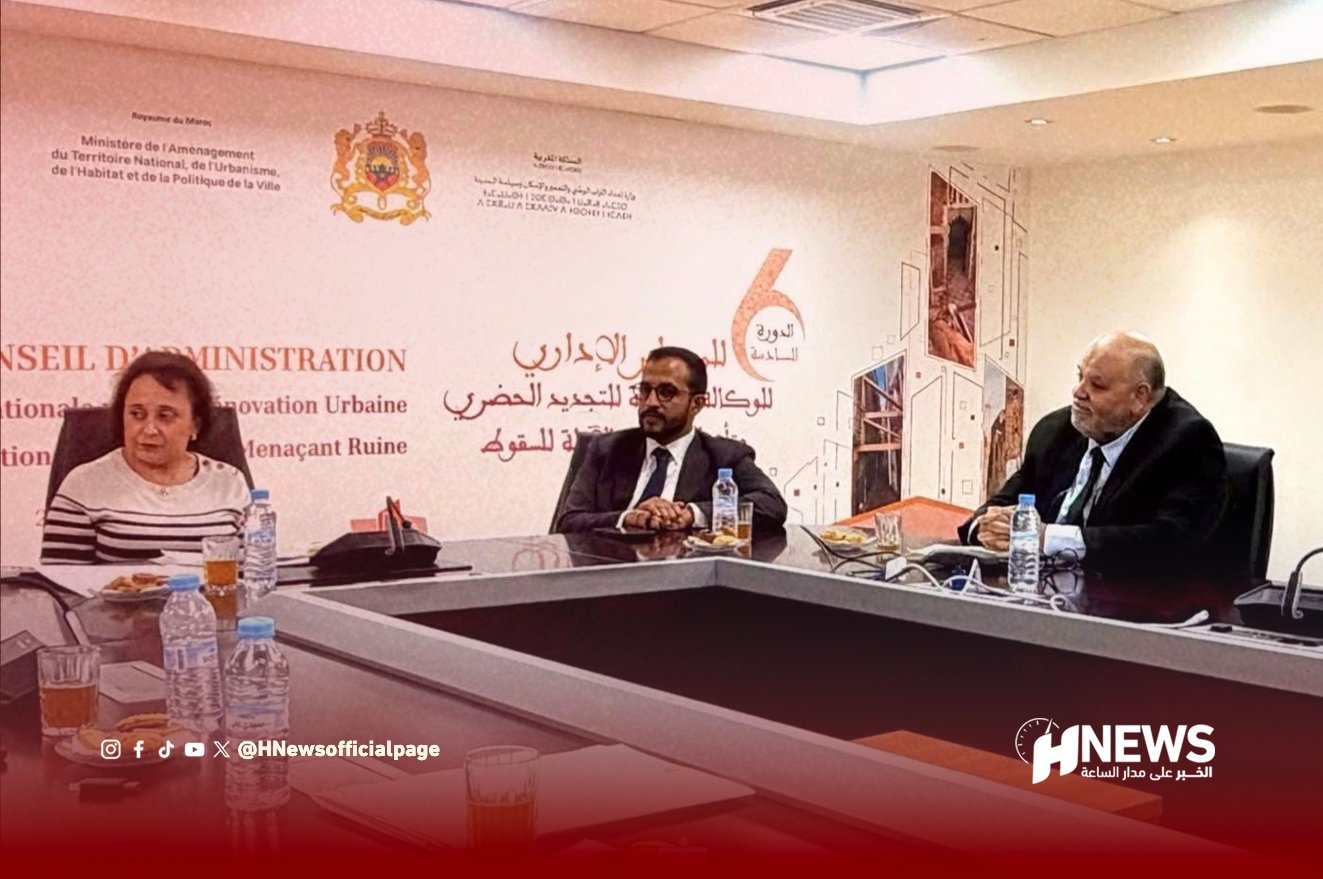Three professionals sit at a long conference table during a panel discussion; backdrop features Arabic text and a crest in a conference room.