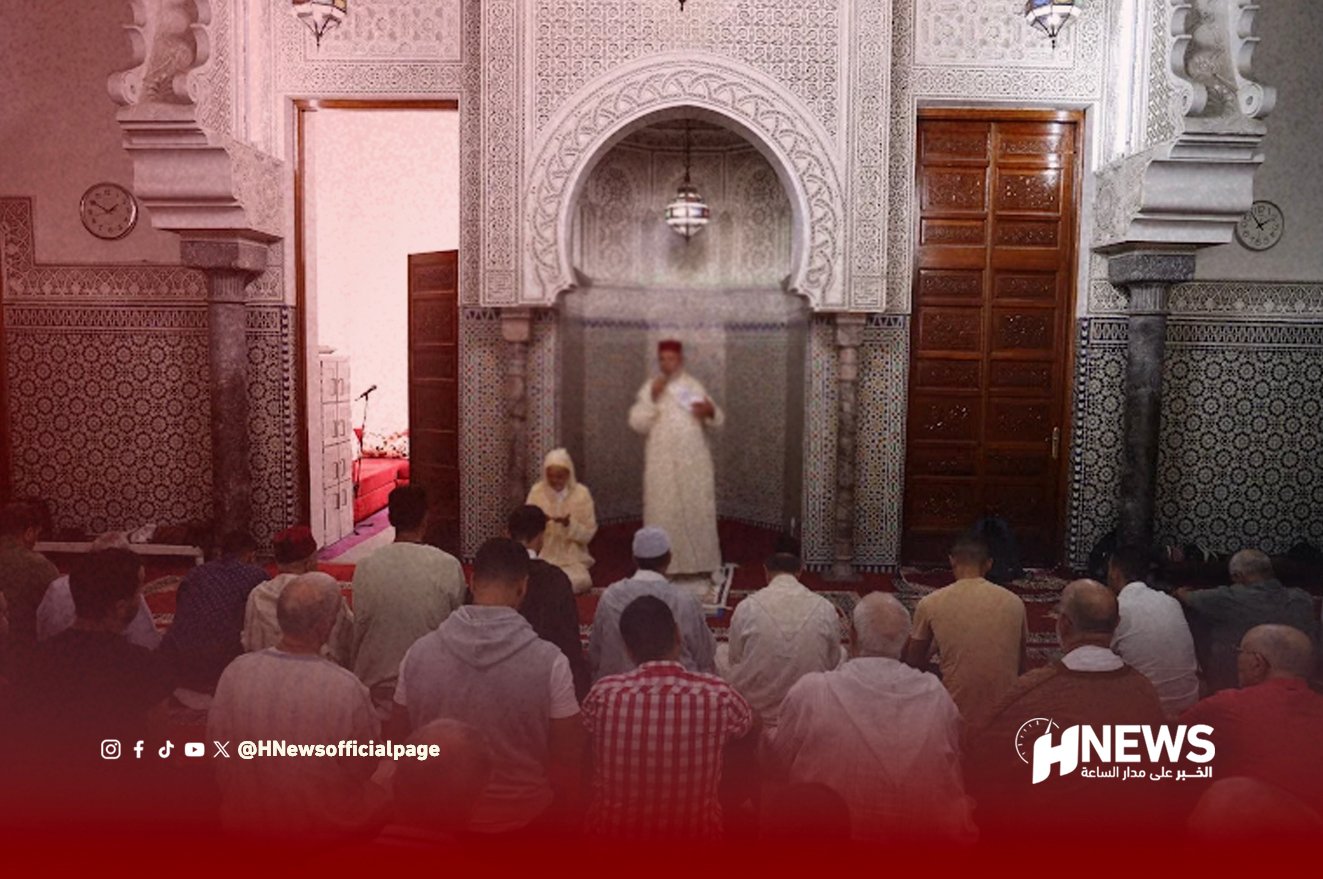 Group of worshippers seated on a red-carpeted floor inside a richly tiled mosque, listening to an imam at the central arch.