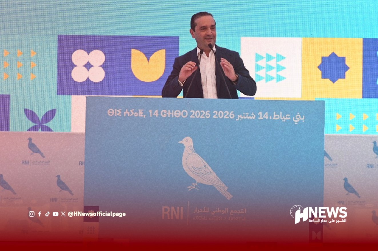 Speaker in a suit delivers a speech at a podium on a blue backdrop with Arabic text and geometric shapes in the background.