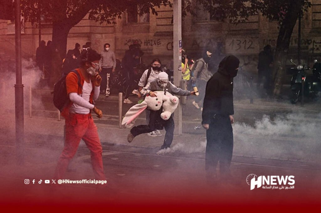 الثورة في فرنسا | H-NEWS آش نيوز Protest scene with smoke on a street; masked demonstrators run, including a person in orange with a backpack and a large white stuffed bunny nearby.