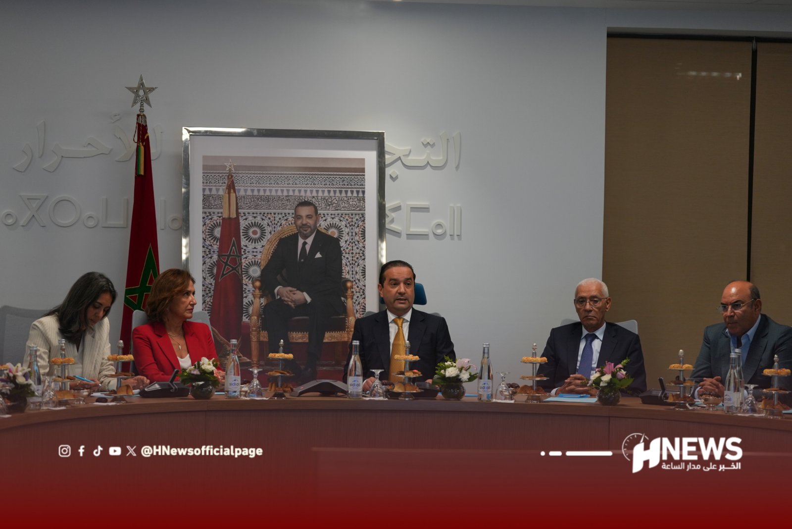 Panel of officials seated at a long conference table with a large framed portrait of a man in a suit and a Moroccan flag behind them.