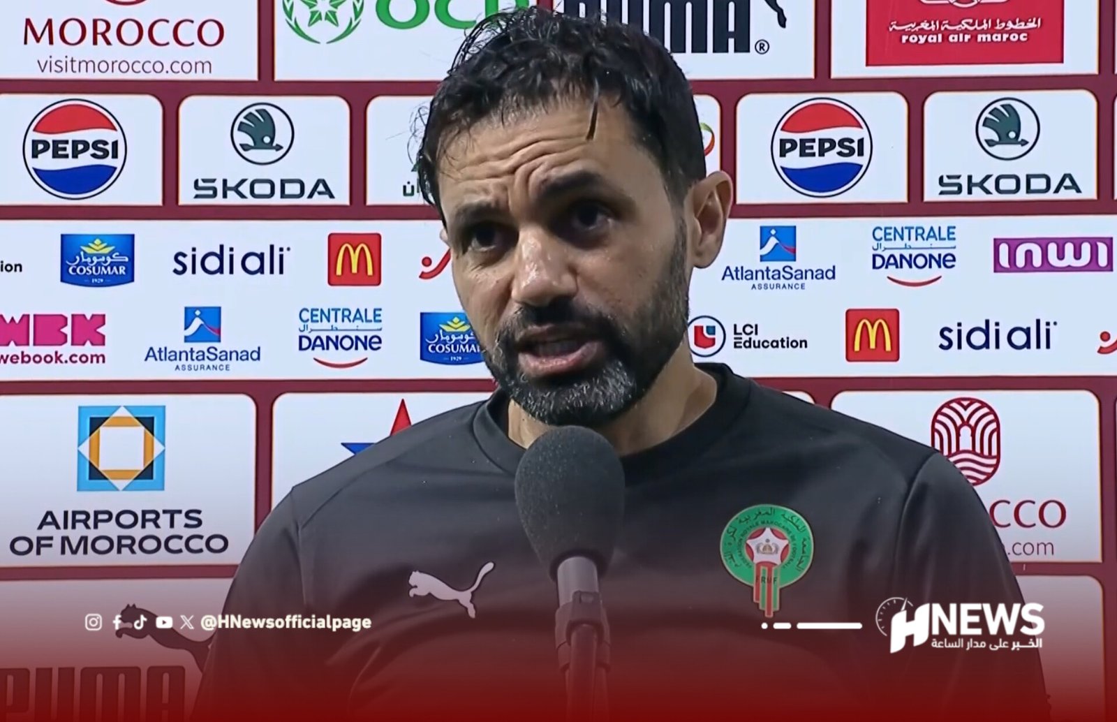 Man speaks into a microphone at a post-match press conference, backdrop covered with sponsor logos and a Moroccan crest on his shirt badge.