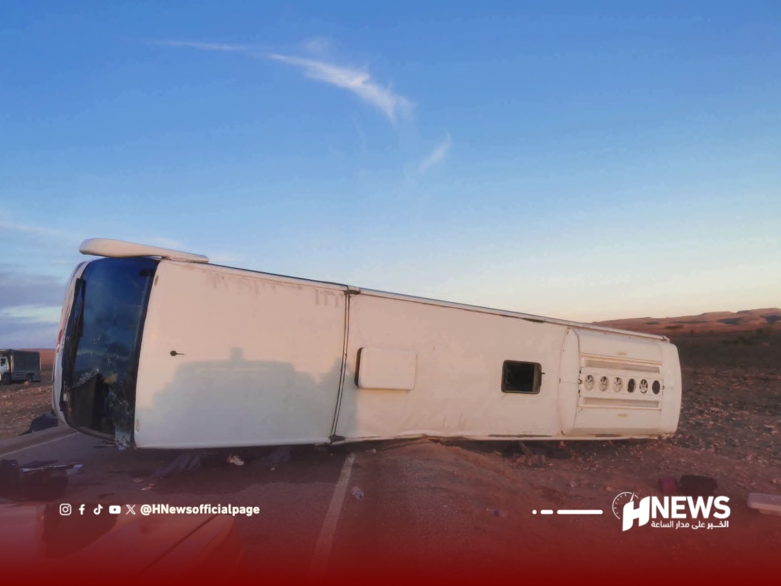 White bus overturned on its side on a desert road beneath a clear blue sky.