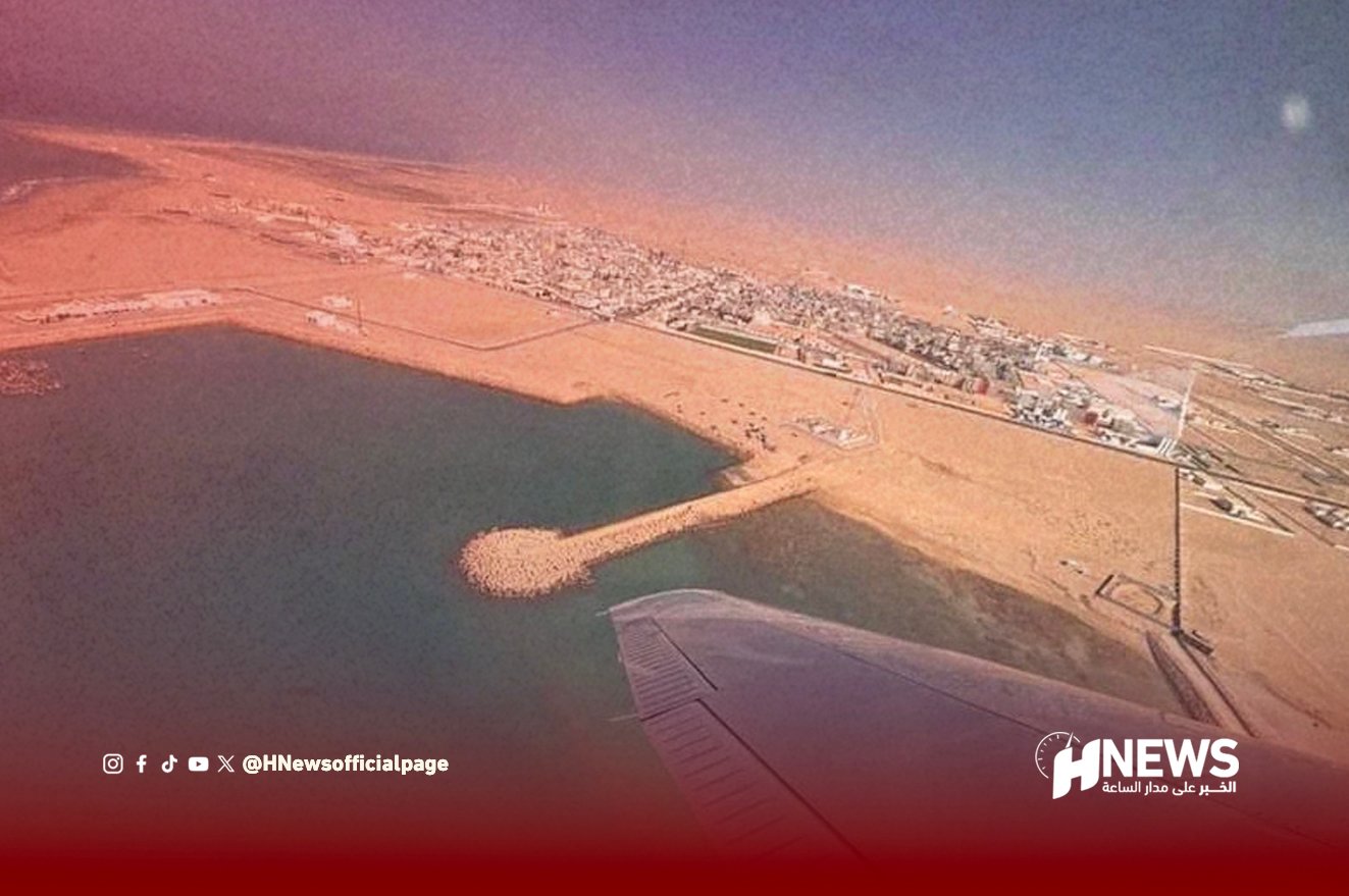 Aerial view from an airplane window of a desert coastal town with a curved breakwater forming a small harbor, visible water and shoreline.
