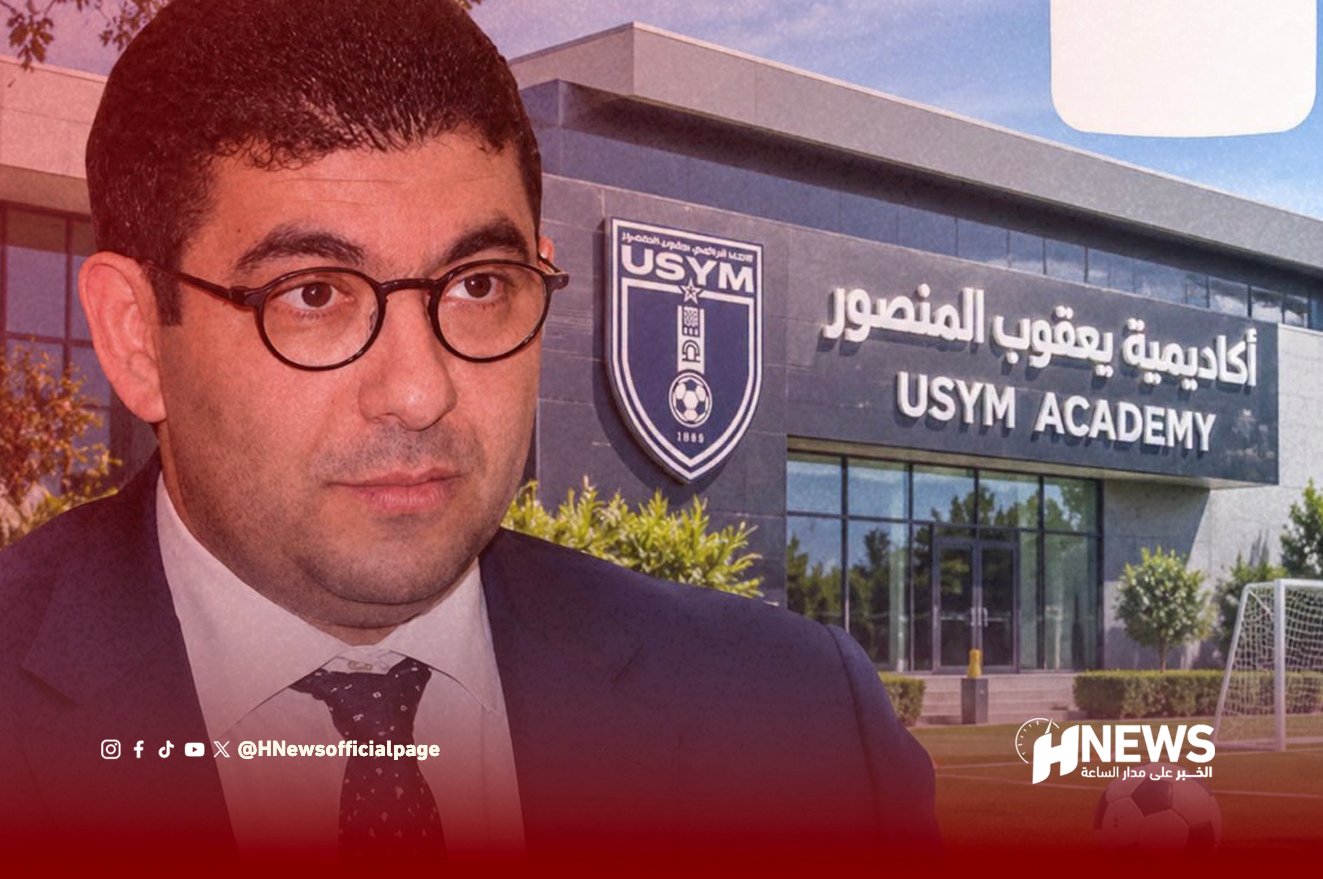 Portrait of a man in a suit and round glasses standing in front of the USYM Academy building.